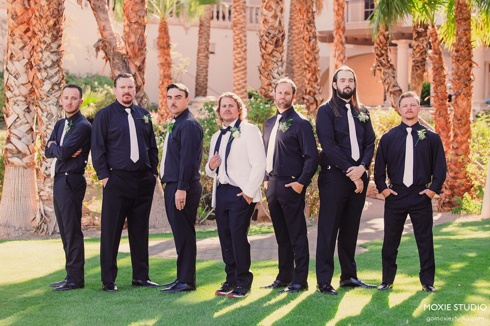 A wedding party poses together in a line outdoors against a backdrop of coral-colored pillars and green foliage.