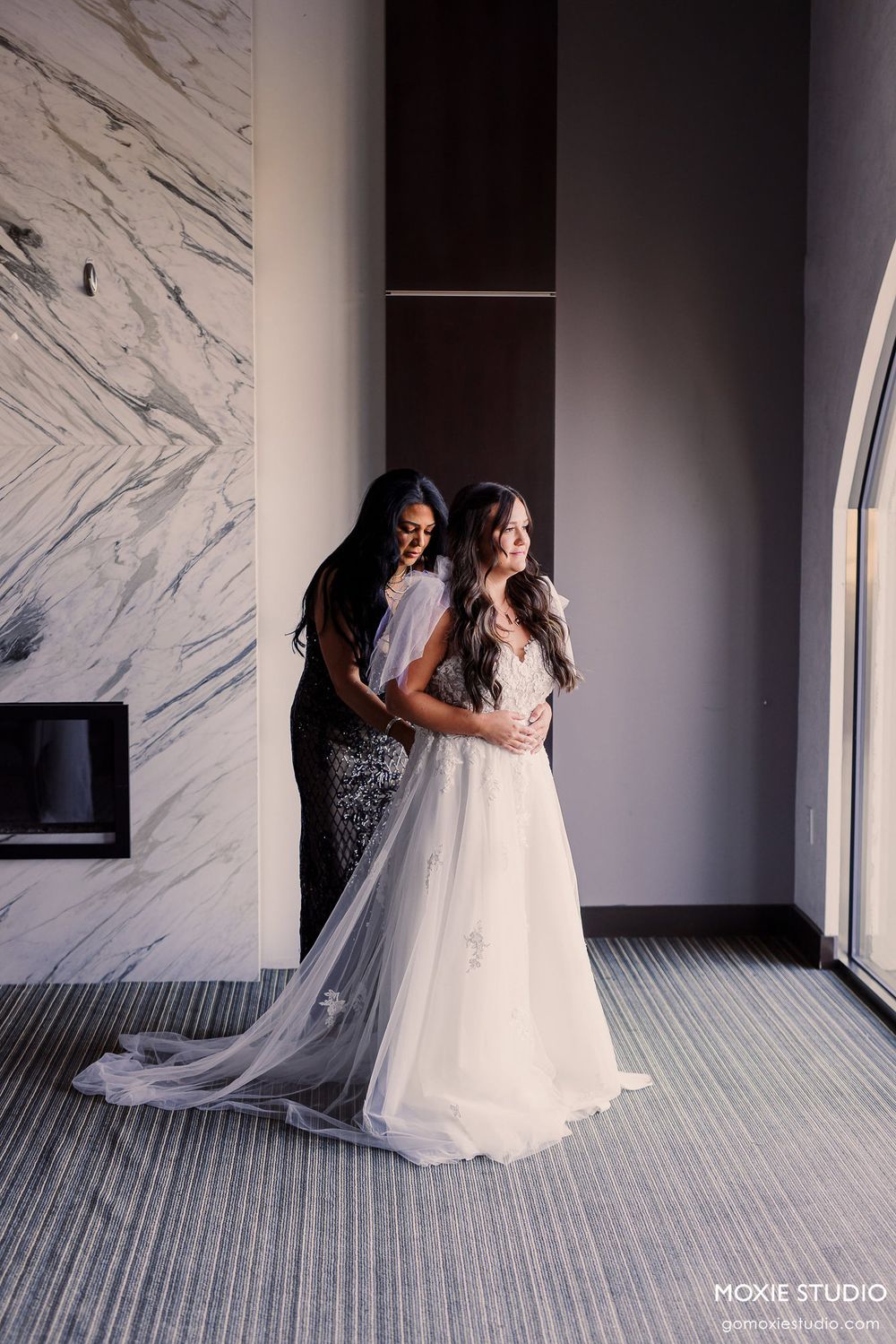 A bride in a flowing white gown stands near a marble wall while getting help with her dress.