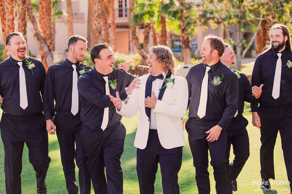 A large wedding party in black and white attire stands in a line outdoors on a sunny day.