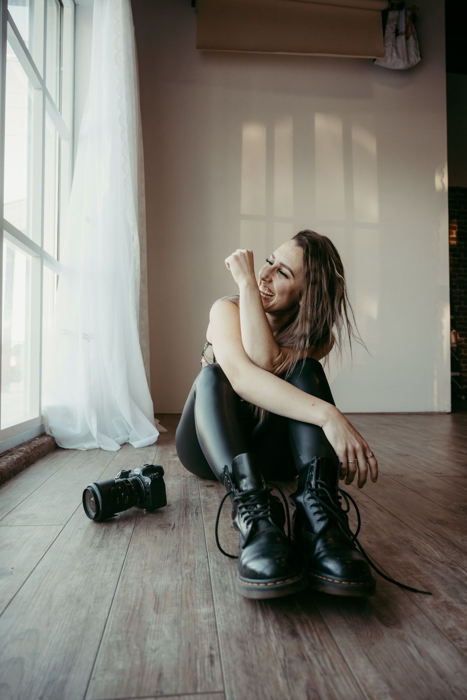 Artist sitting on wooden floor near window with camera, wearing combat boots in moody lighting.