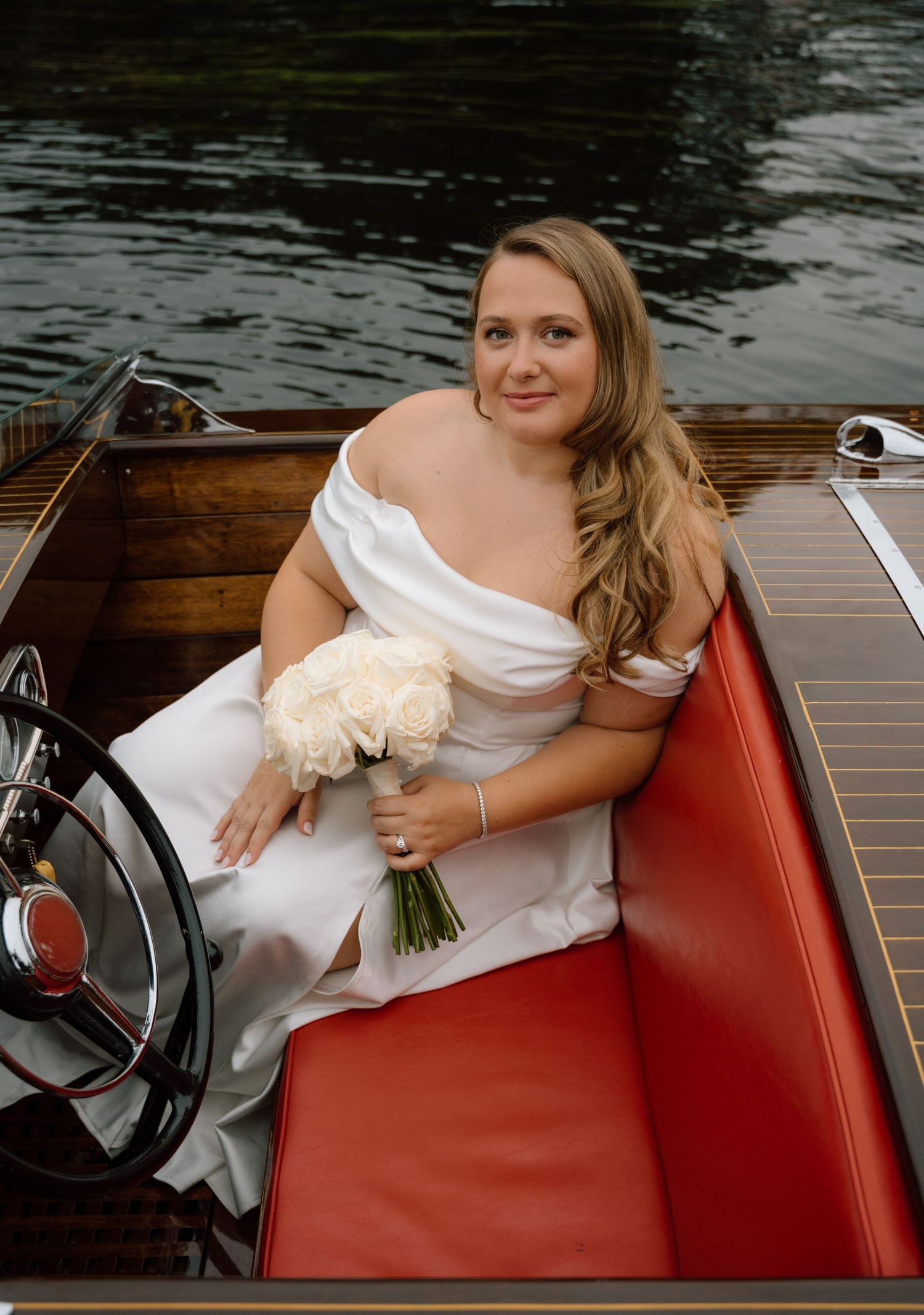 A wedding couple sits in a wooden boat with flowing white veil trailing behind them on the water.