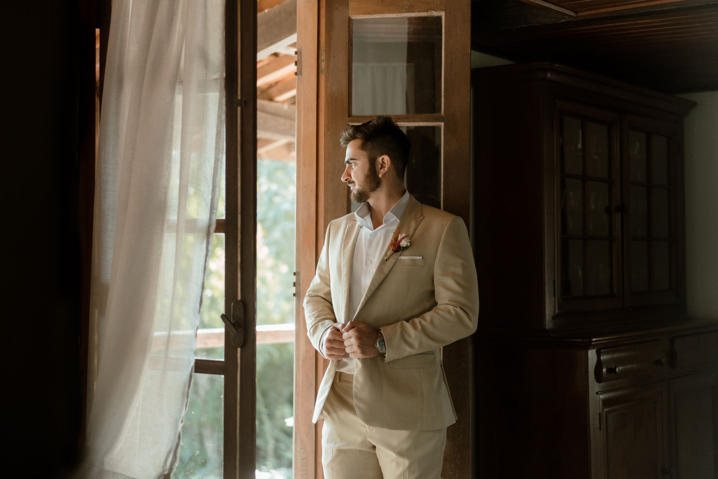 Figure standing by window in beige suit gazing at natural light streaming through sheer curtains.