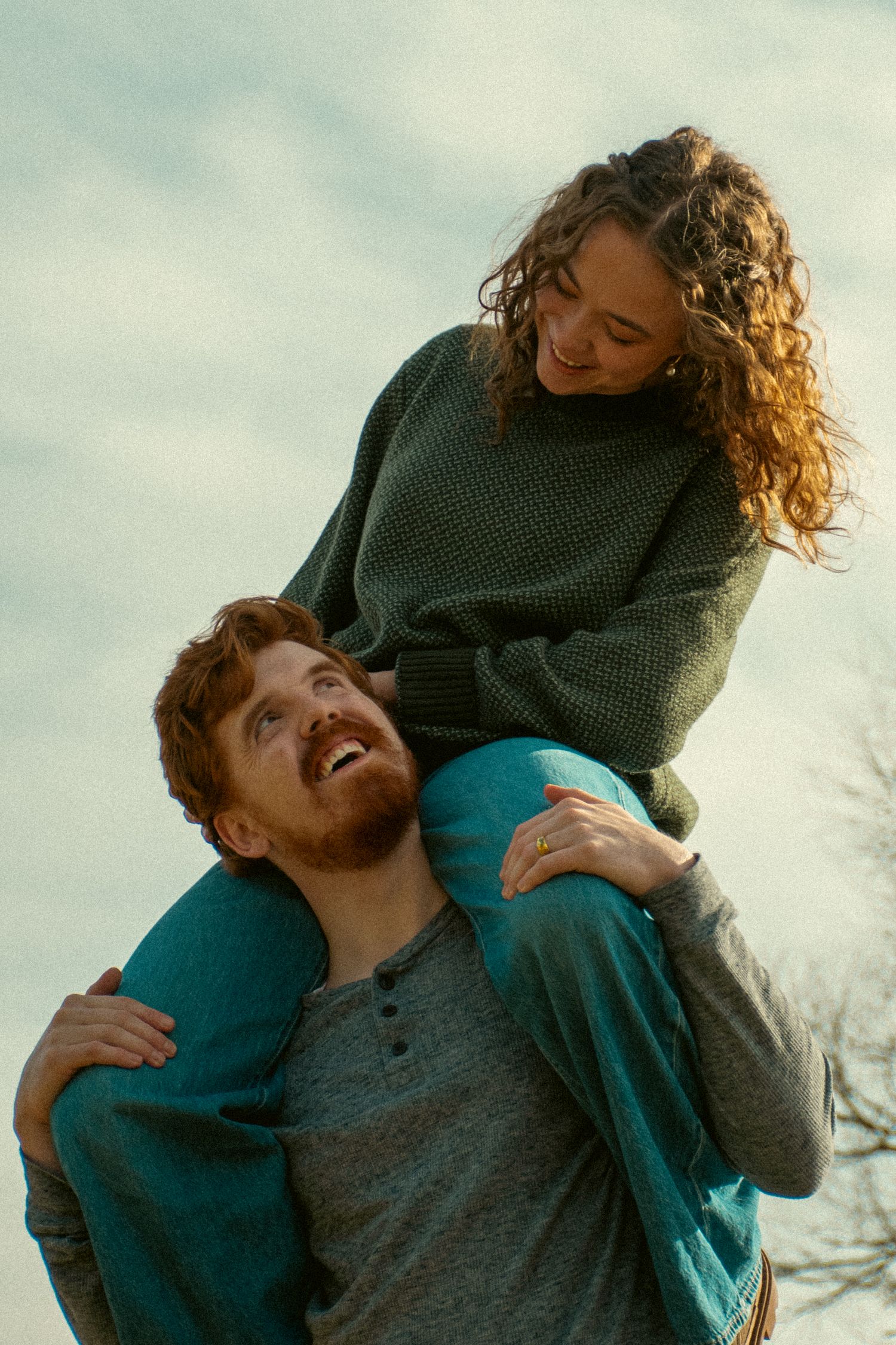 A couple shares a joyful cinematic moment outdoors as one carries the other on their shoulders against a soft sky backdrop in Draper Utah.