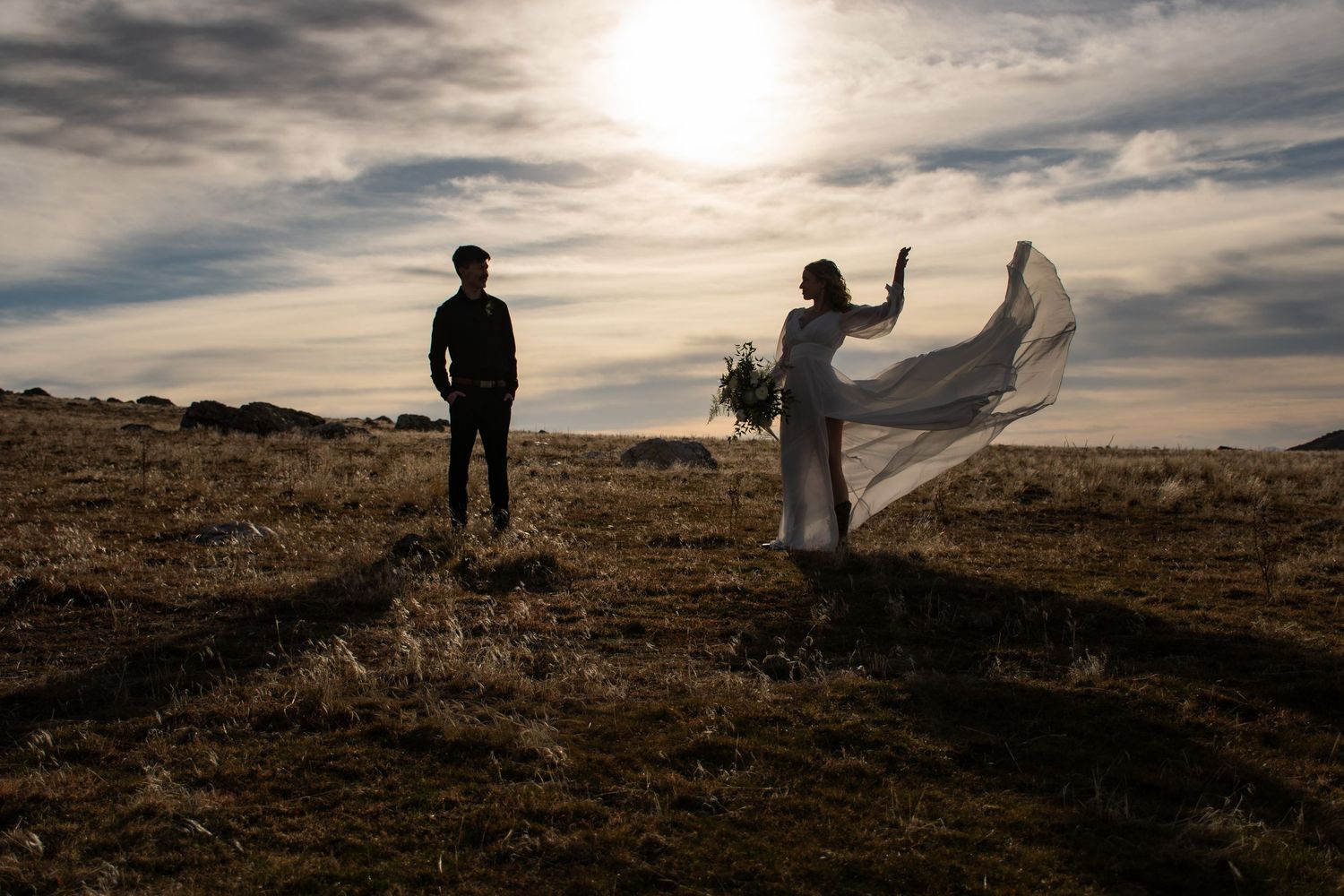 A silhouetted couple poses dramatically on a hilltop with flowing dress and moody sky at Antelope island in Utah.