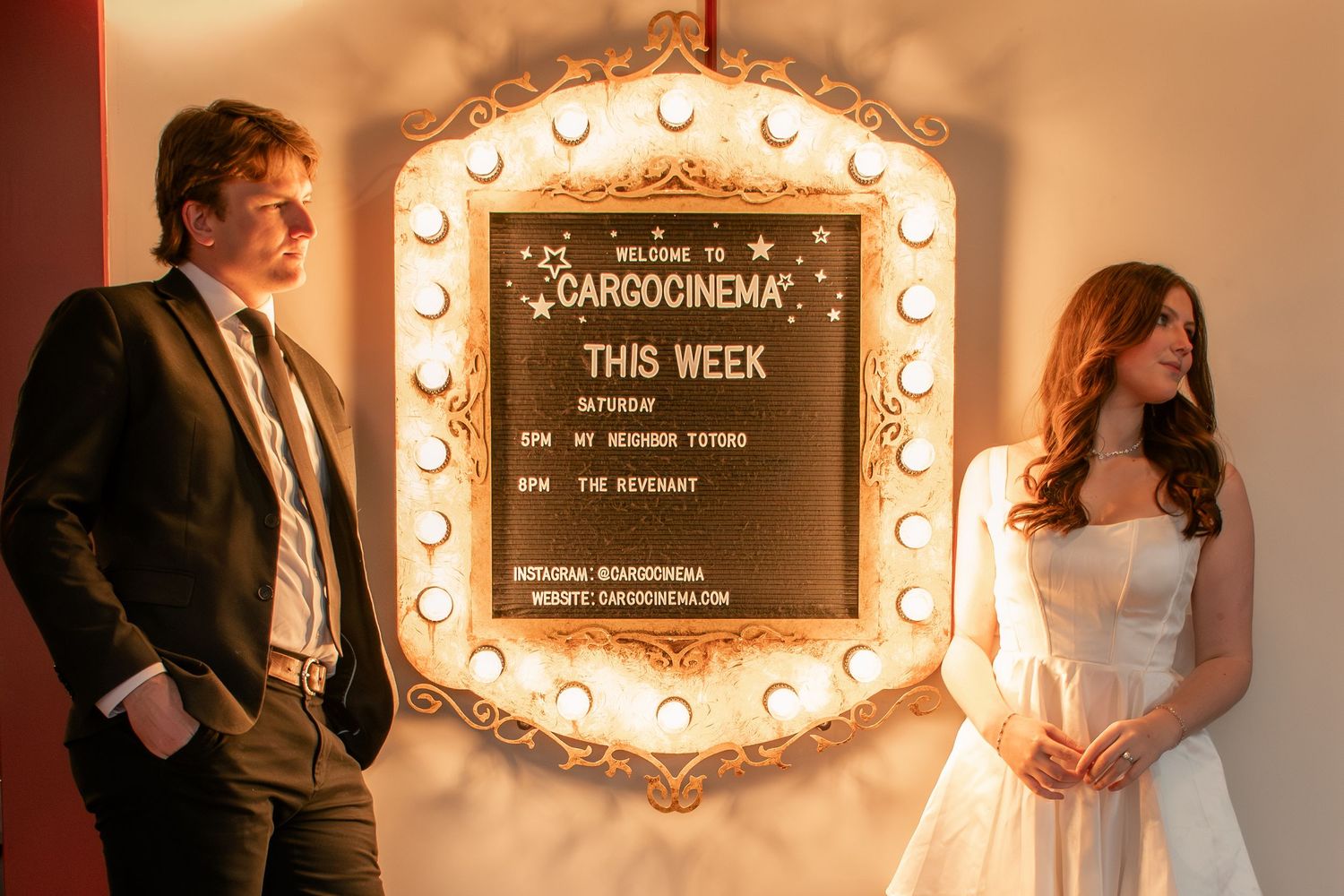 A couple poses by an illuminated vintage marquee sign with glowing bulbs, in a  cinematic photo at a theatre in Utah.