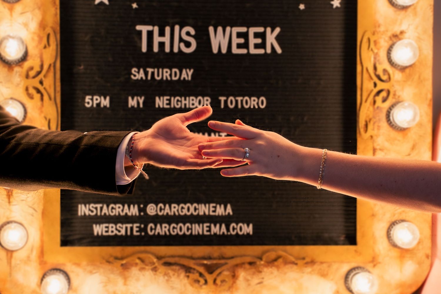 Two hands reach toward each other in front of an illuminated movie showtime board, in a cinematic photos of hands in A theatre in Utah.
