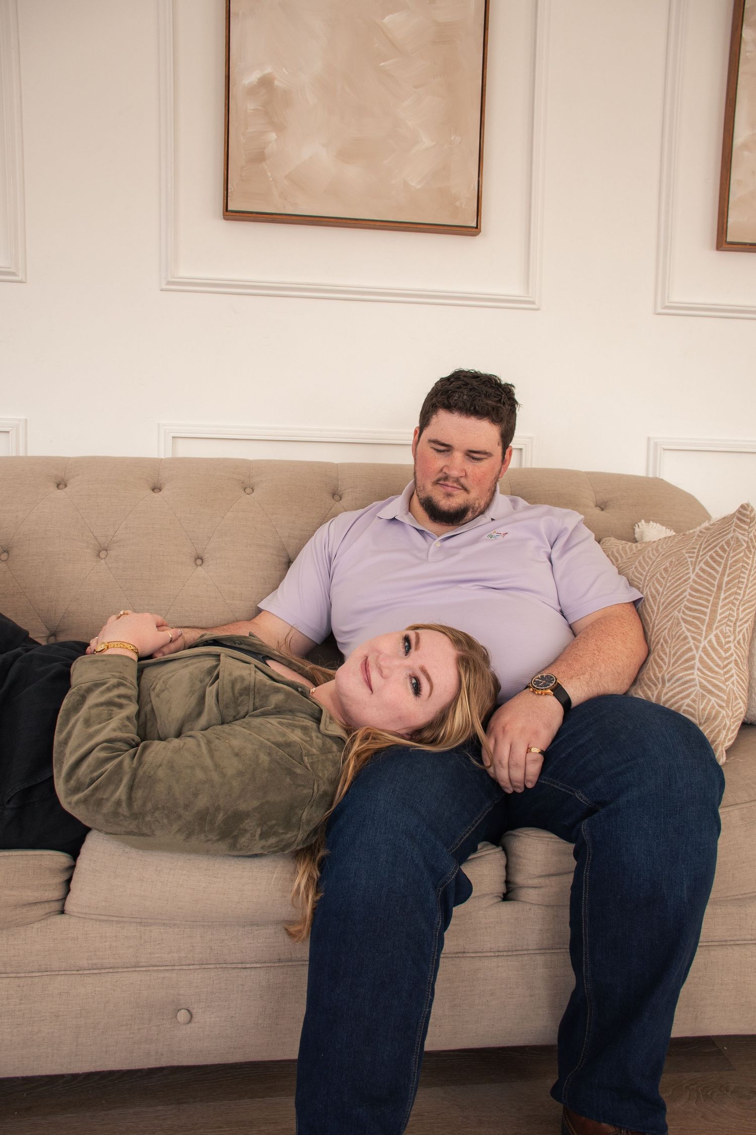 A couple relaxes together on a beige tufted couch in a room with white wainscoting walls.