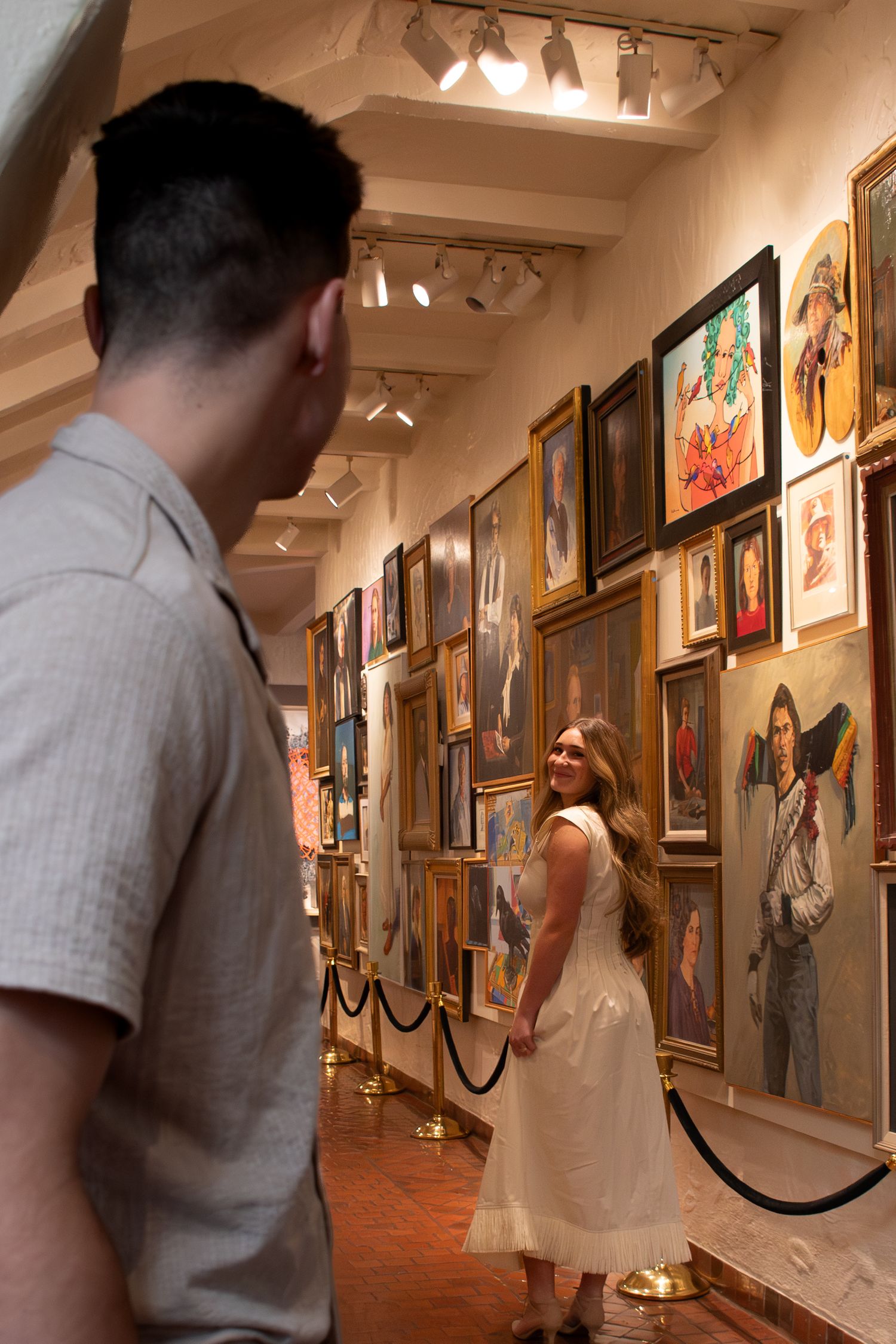 Art gallery visitors admire paintings and artwork displayed along a warmly lit hallway with red carpeting.