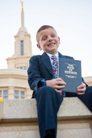 Cute 8-year-old boy grins in front of temple while holding scriptures for his baptism photos.