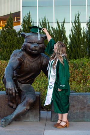 UVU student wearing gown playfully puts cap on mascot statue.