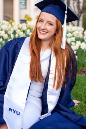 BYU student wearing cap and gown smiles in front of flowers.