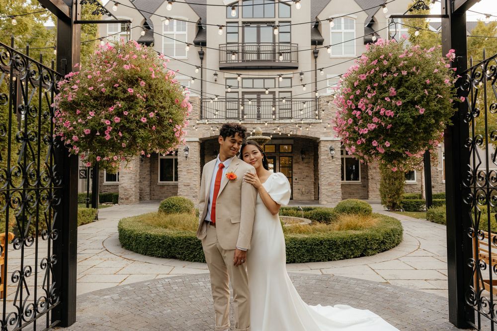 A bride and groom pose under a floral arch at an elegant stone building venue.