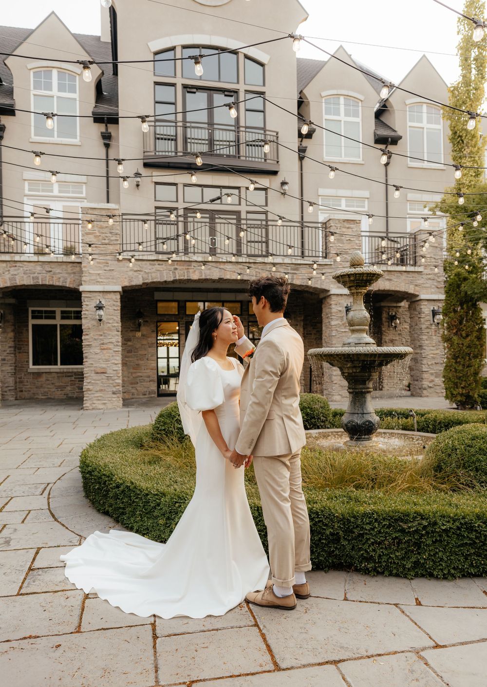 Bride and groom hold hands smiling before a grand stone manor with fountain and string lights.