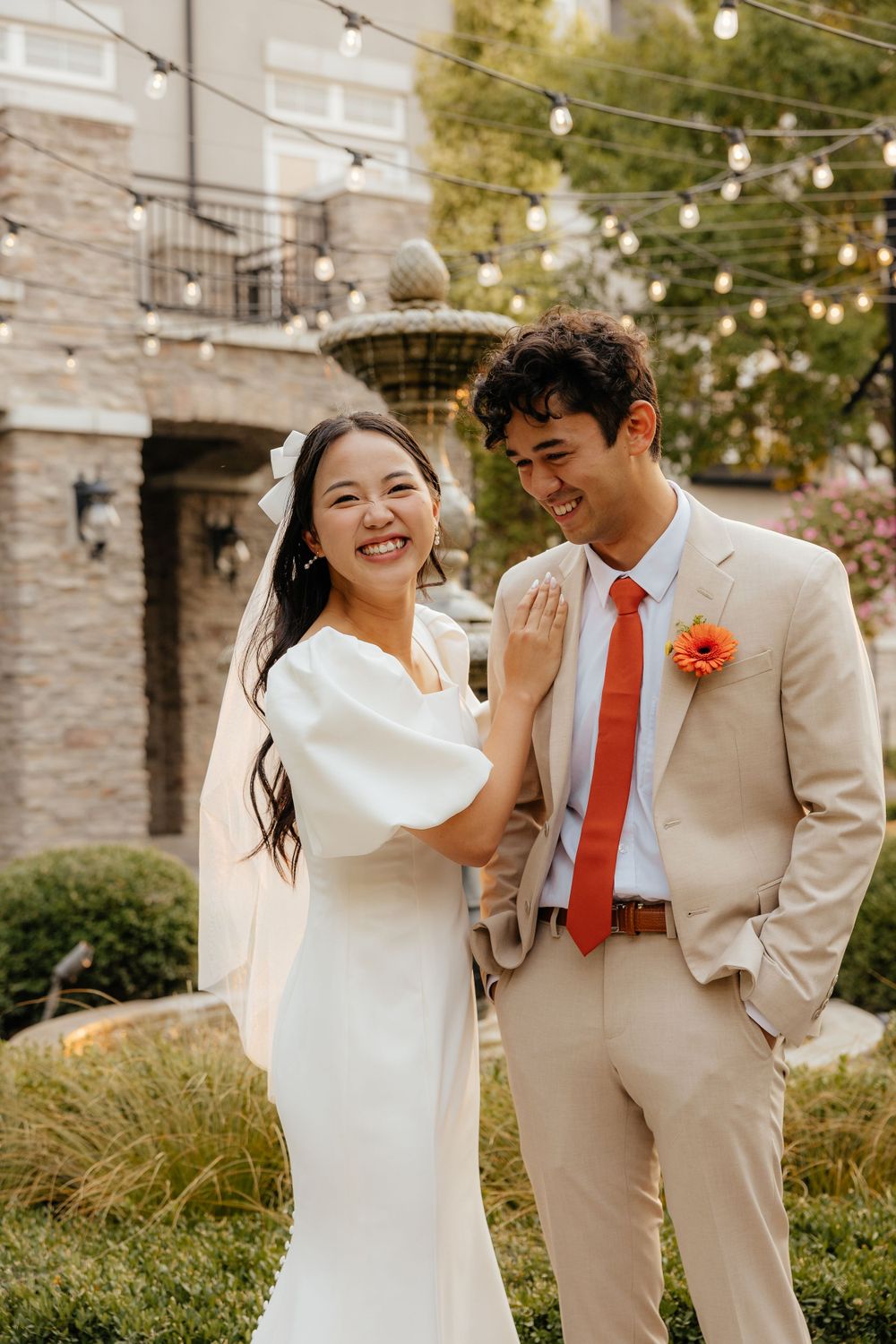Smiling bride and groom in beige suit with orange tie posing in garden venue.