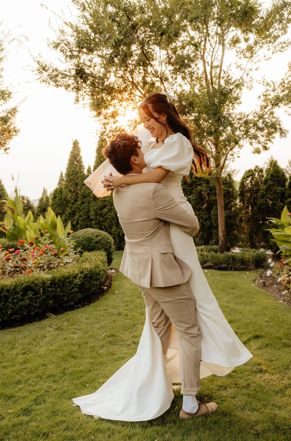 Groom lifting bride in white puff-sleeve gown in lush garden at golden hour.
