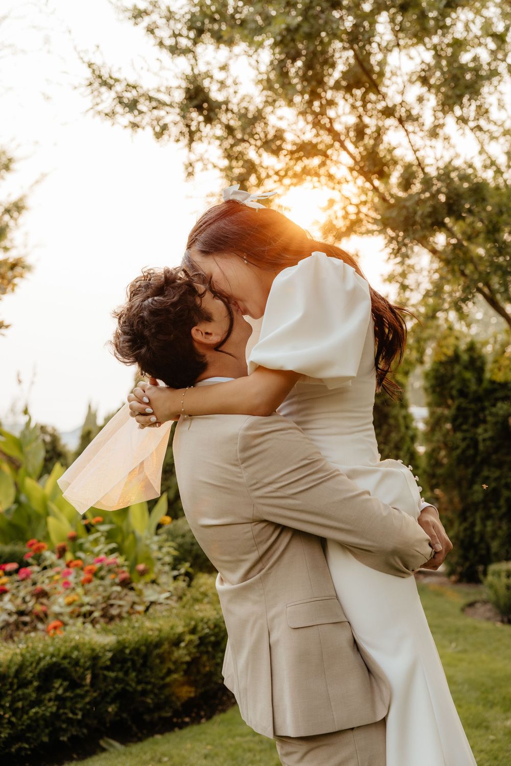 Groom lifting bride in white dress during romantic sunset wedding photo.