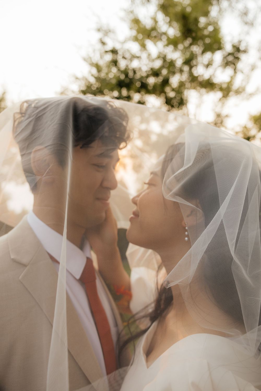 Romantic bride and groom sharing intimate moment under bridal veil at sunset.