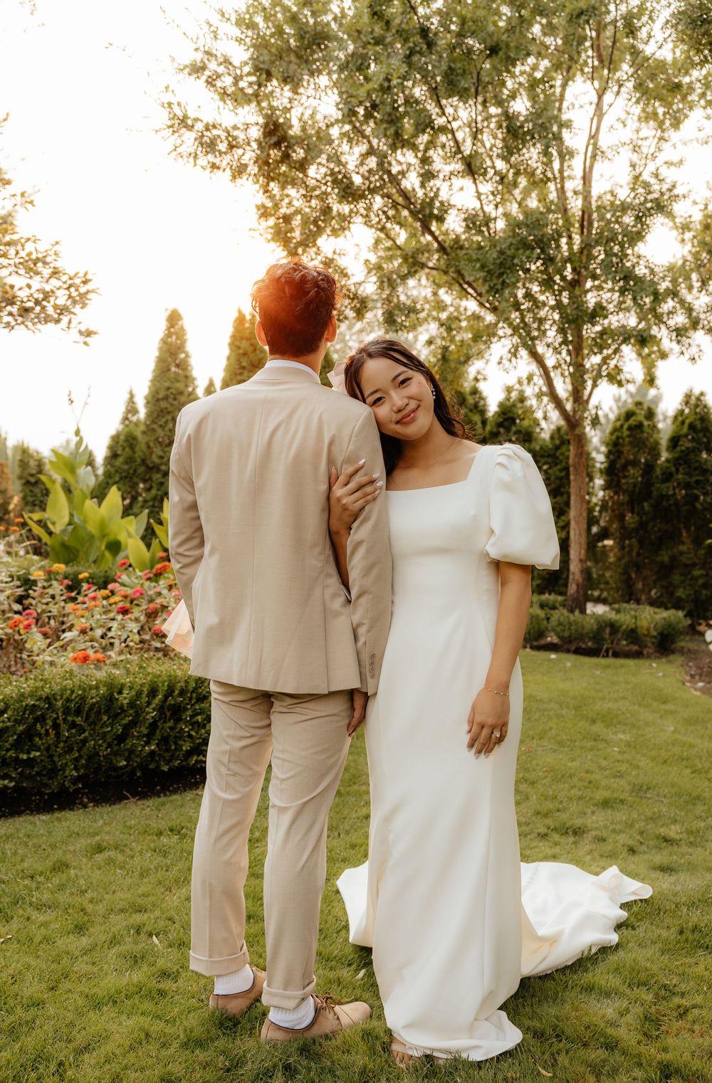 Bride in white puff-sleeve gown smiling beside groom in beige suit in garden.