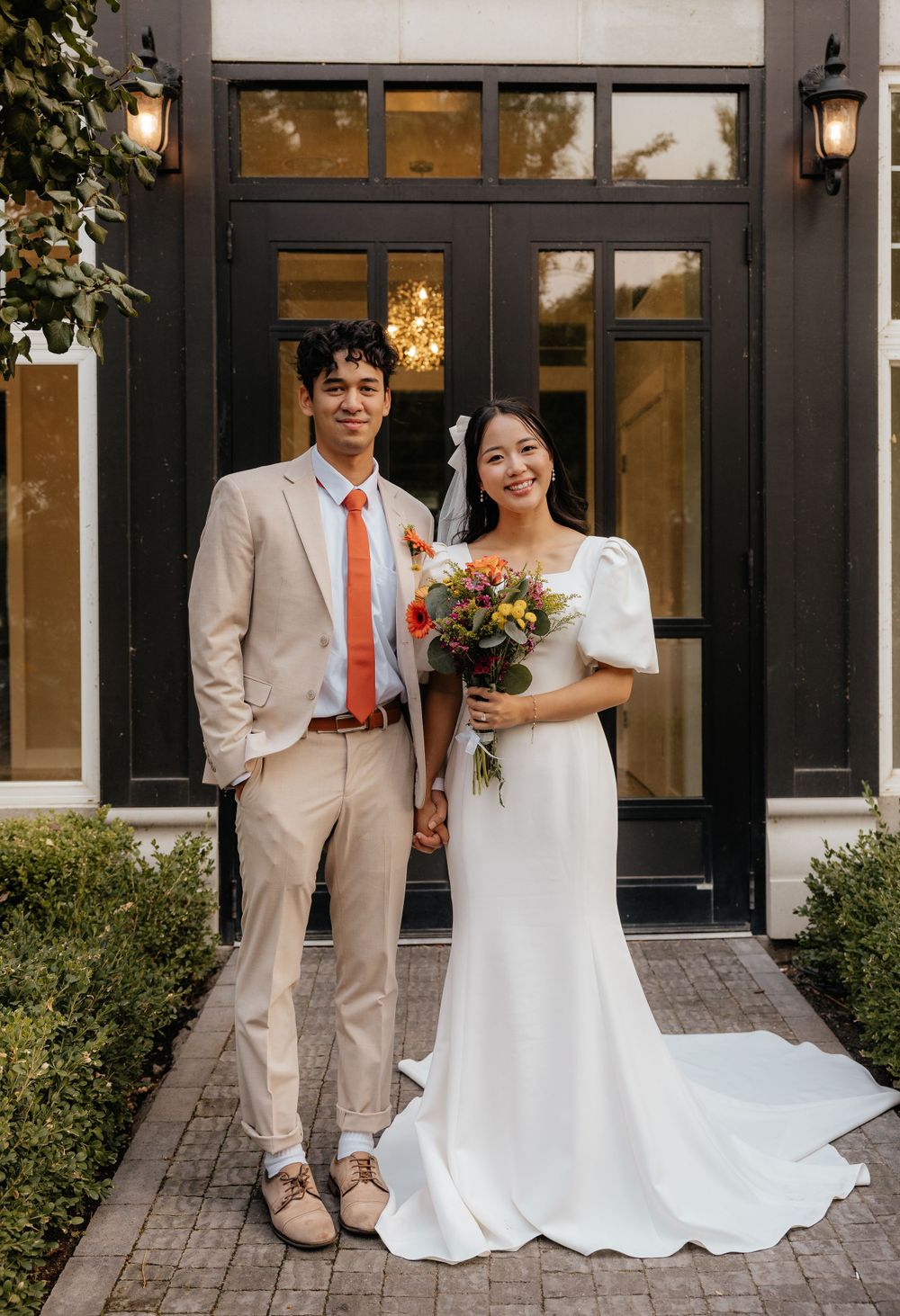Bride holding colorful bouquet and groom in beige suit posing at venue entrance.