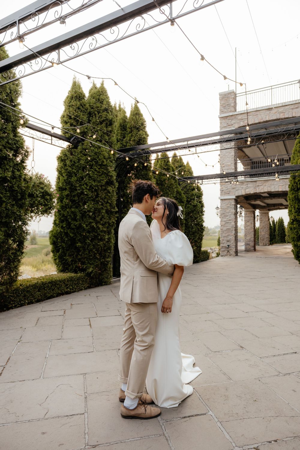Bride and groom share a tender kiss under a pergola with string lights and tall cypress trees.