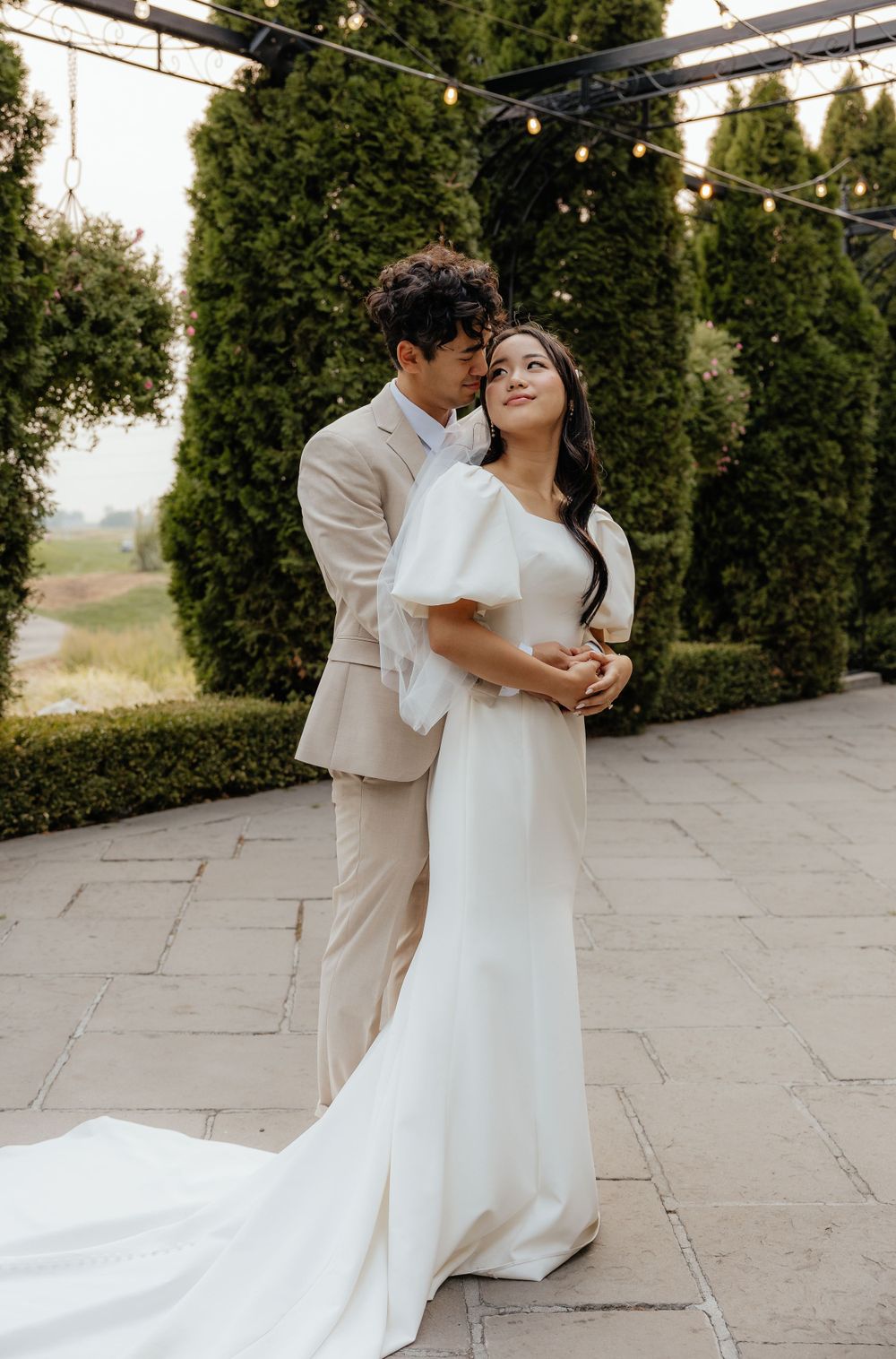 Bride and groom share a romantic embrace on a stone patio surrounded by tall evergreen trees with string lights.