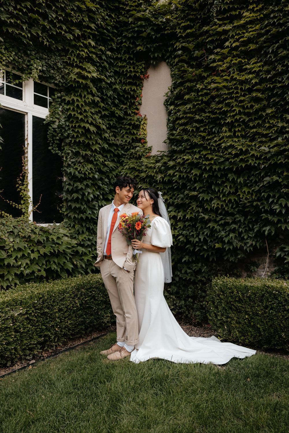 Wedding couple stands before an ivy-covered wall, bride holding vibrant orange bouquet in white dress.