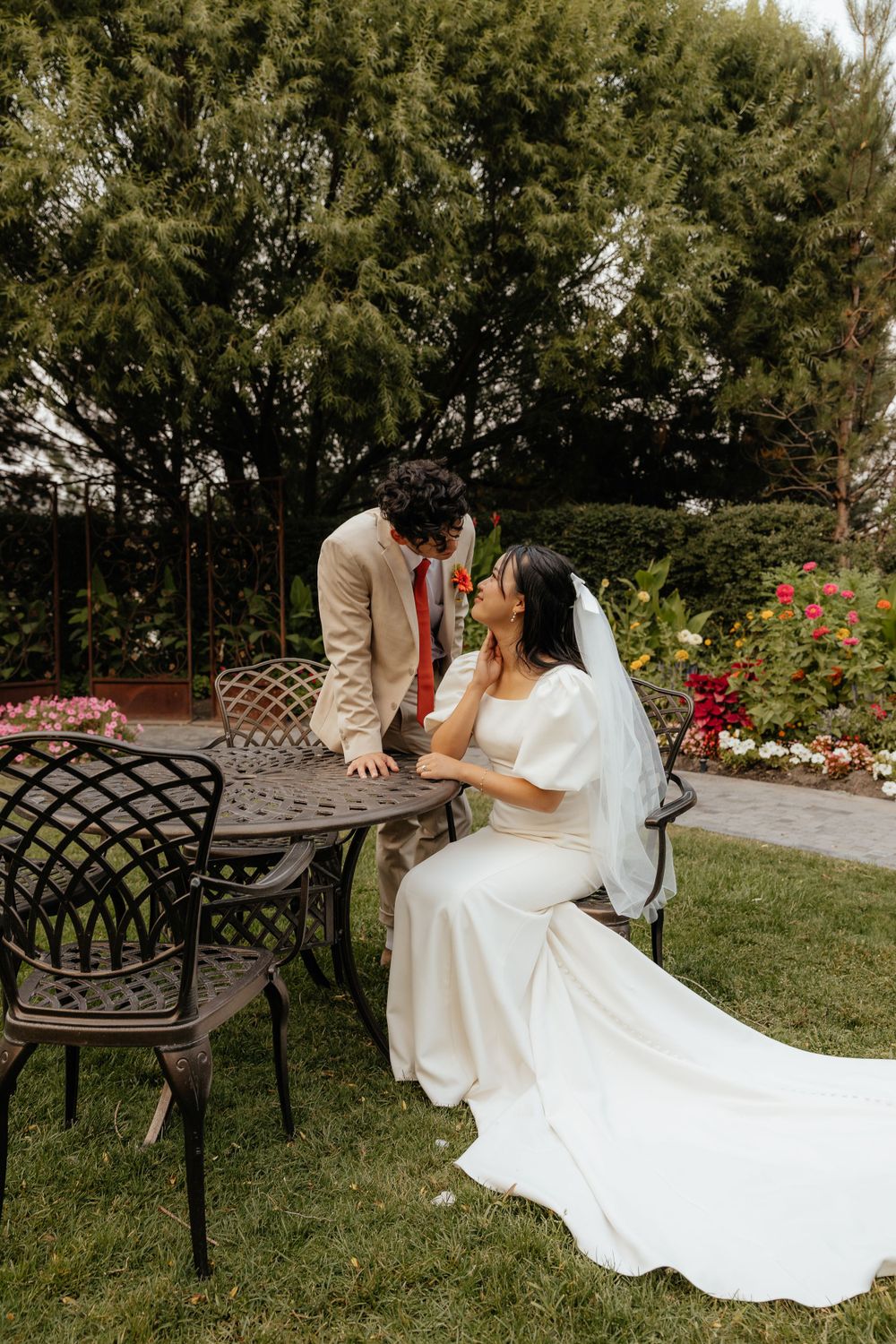 Bride in white gown sits at ornate garden table as groom leans in for a kiss amid colorful flowers.
