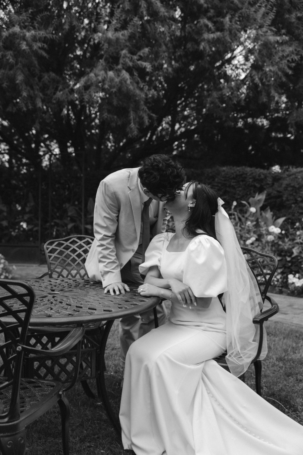 Black and white photo of bride and groom sharing a kiss at an elegant outdoor garden table.