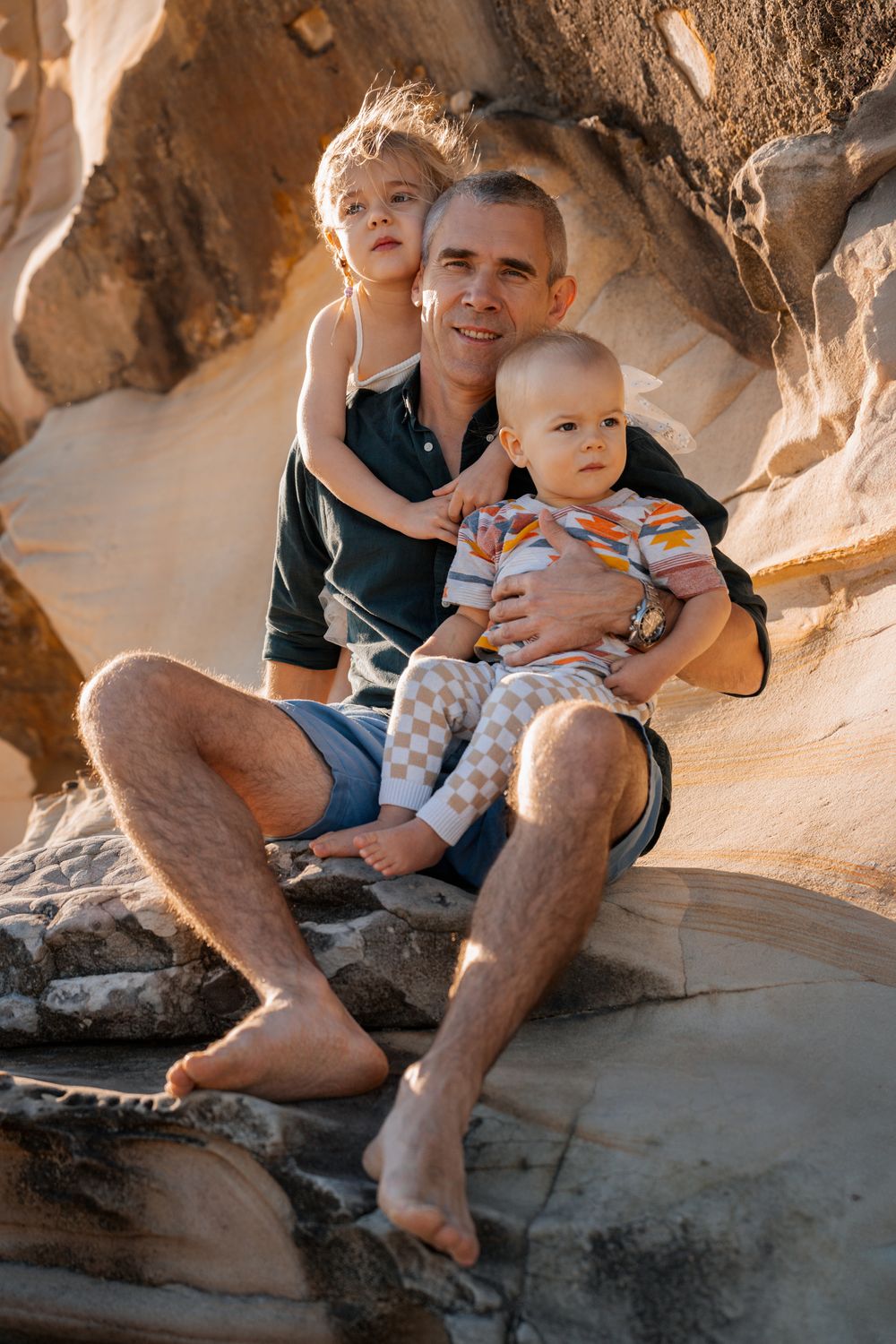 dad sits on the rocks with his daughter and son during a family photoshoot at north curl curl sydney