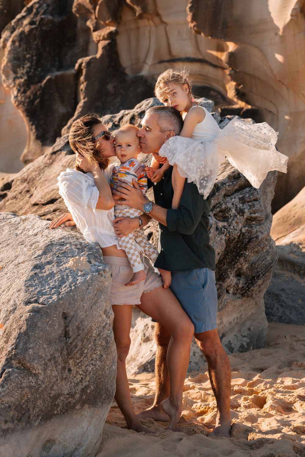 dad and mom kissing toddler while daughter sits on dad's shoulder during a golden hour at the beach family photoshoot