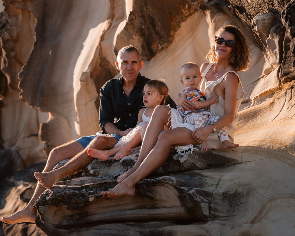 family of four sits on the rocks and looking at the family photographer in sydney during a golden hour time at curl curl