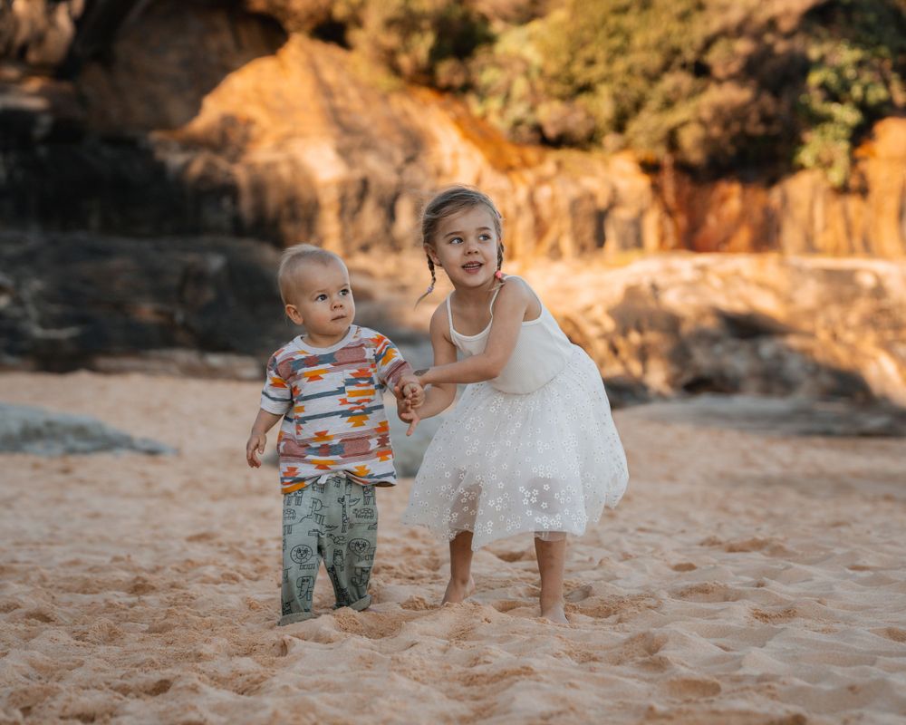 girl and boy siblings are holding hands and looking out of the pictures watching mom during a family photoshoot in sydne
