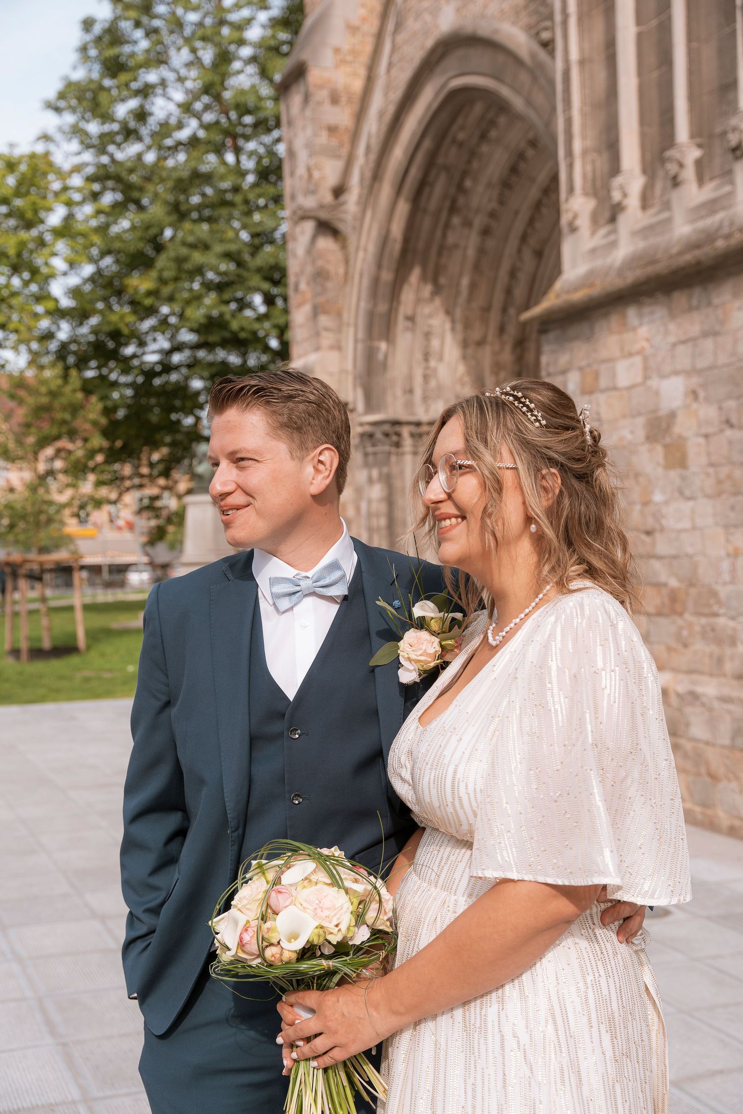 A joyful wedding couple poses together outside a historic cathedral with spring flowers.