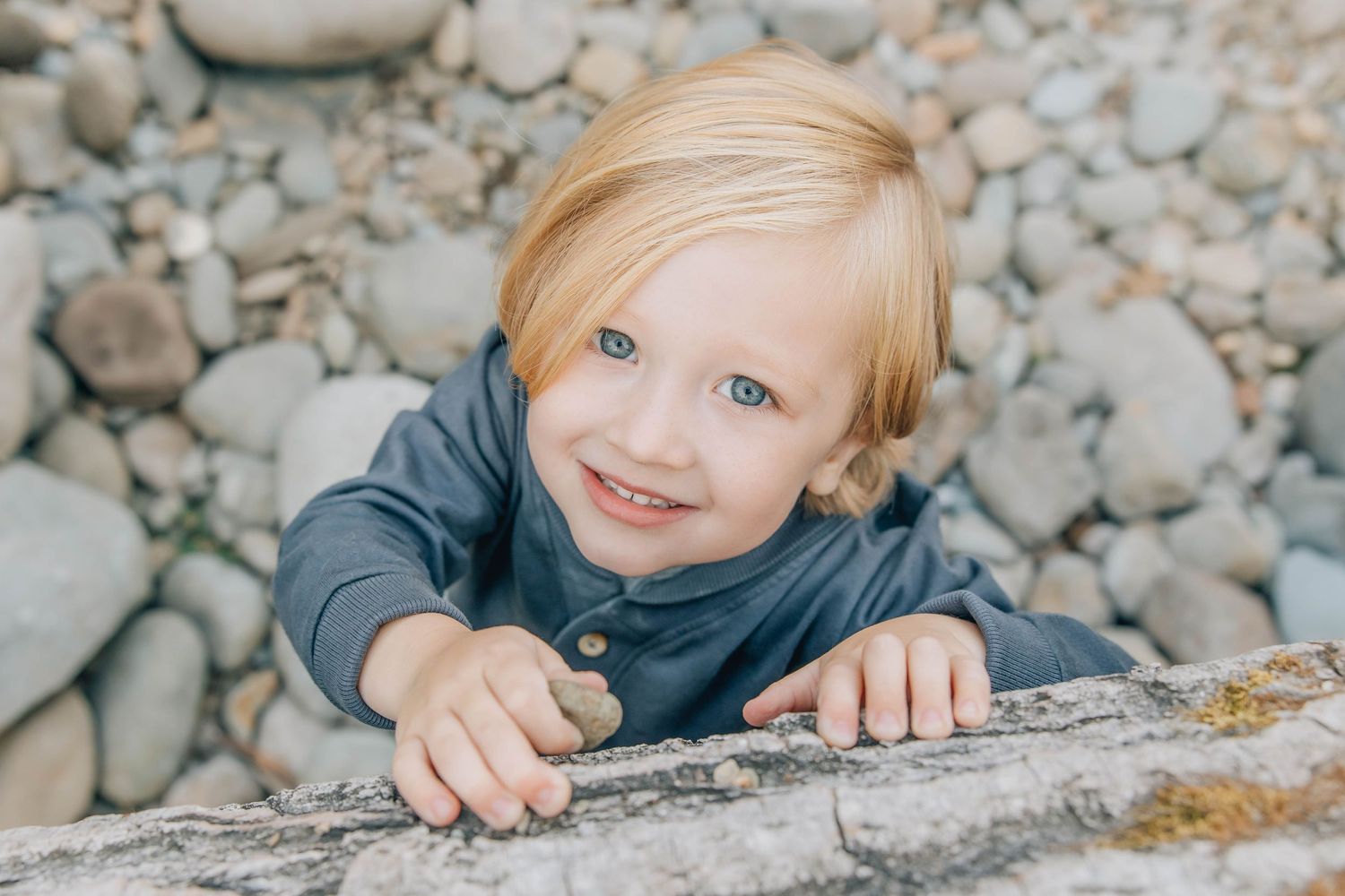 Smiling toddler with blonde hair and blue eyes leaning on a rock outdoors.