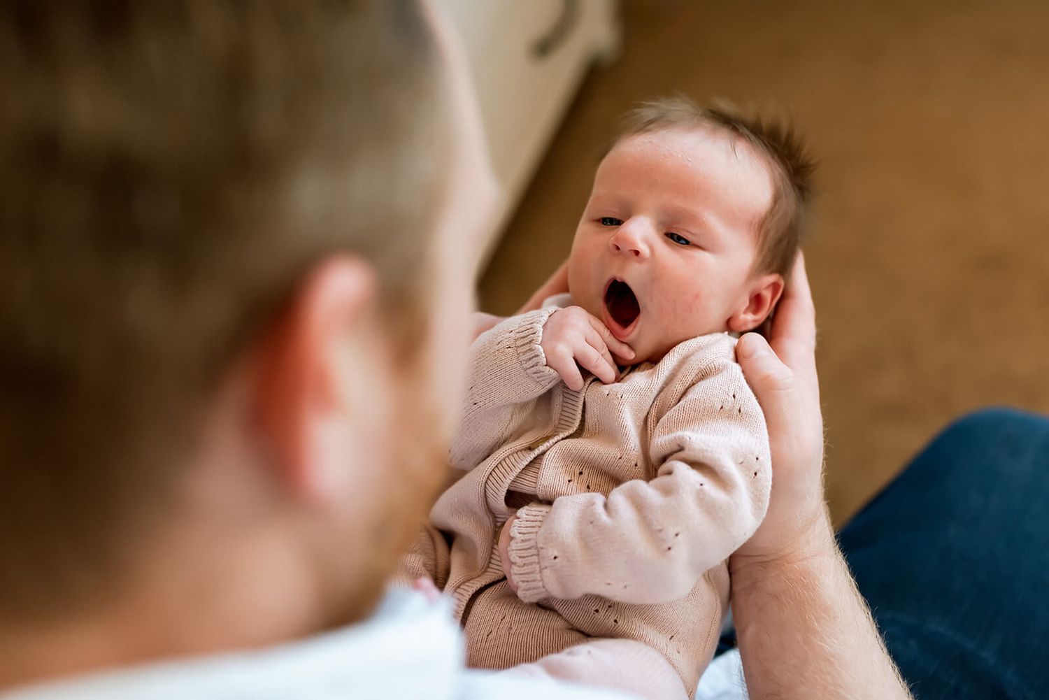 Newborn baby yawns while being held in white onesie.