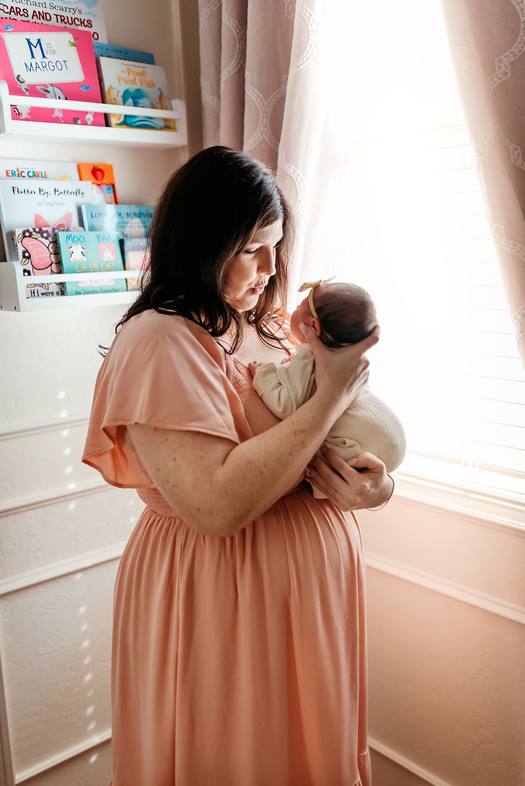 A tender nursery scene shows a parent in a peach dress cradling a newborn by a window with children's books on shelves.
