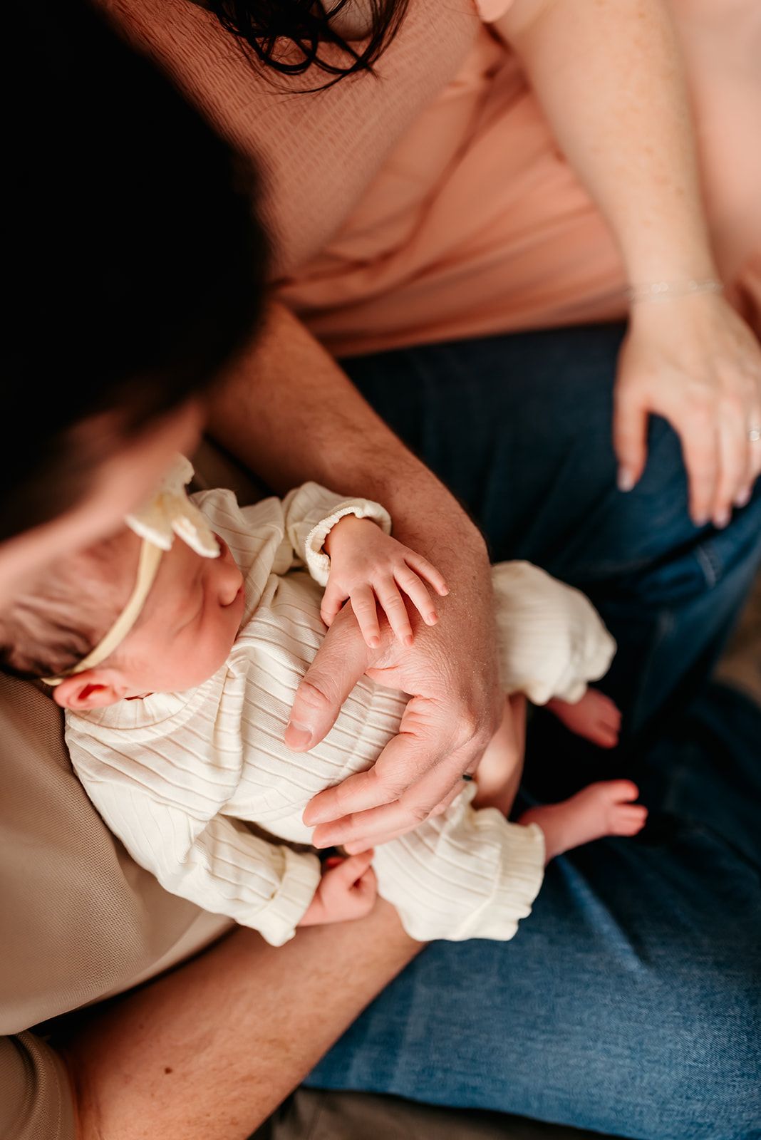An infant wearing a white knit outfit rests in someone's lap while their hands gently hold the baby's small hands.