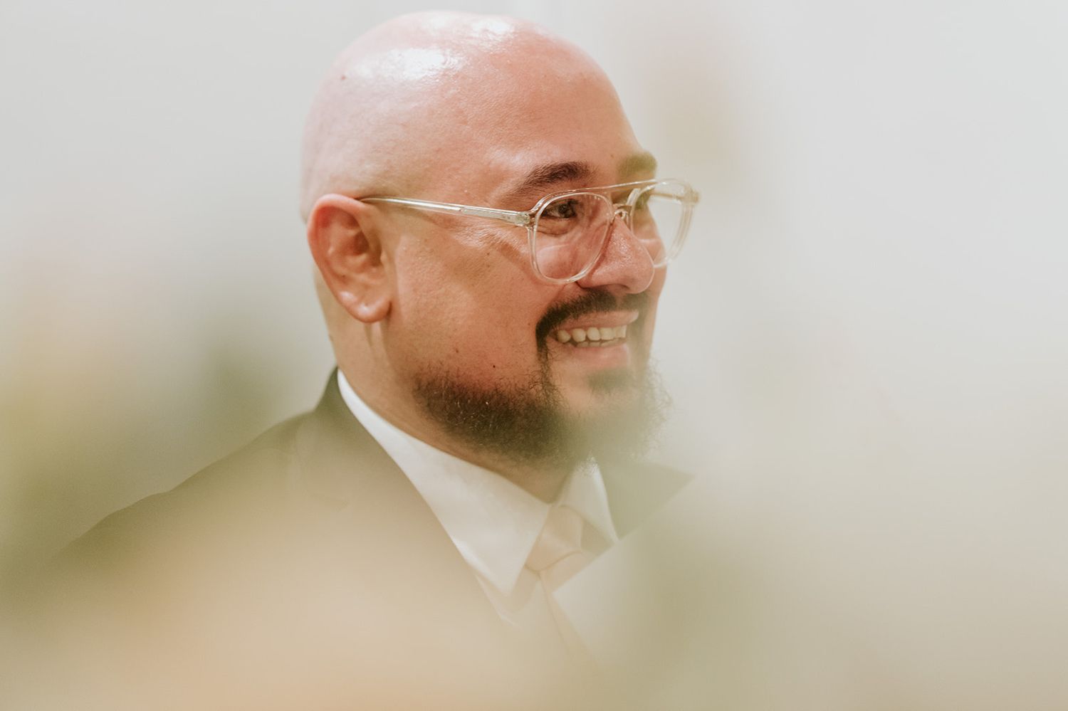 A joyful man in formal attire with glasses and beard photographed through a soft focus lens.