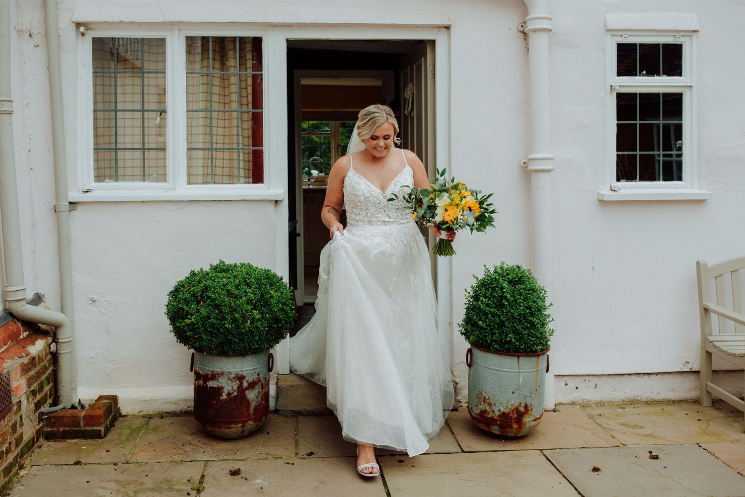 Bride in a white wedding dress holds yellow flowers while standing outside a house entrance. Bride entrance houchins