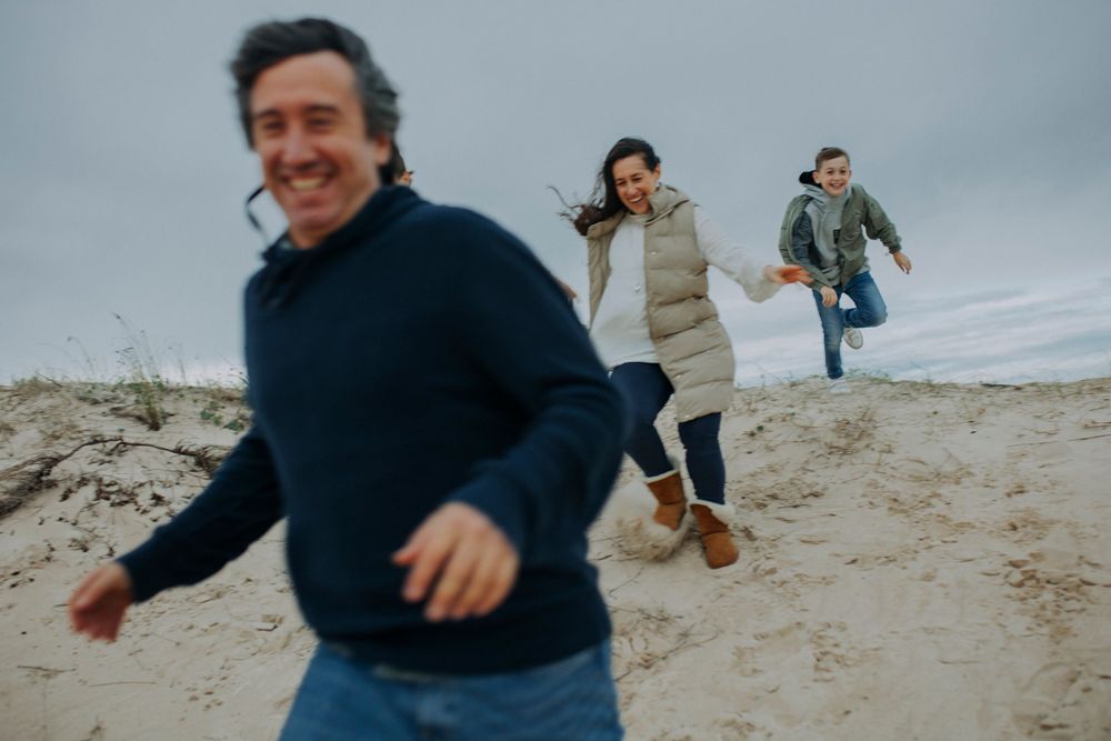 Three people running and playing together on sandy beach under cloudy sky.