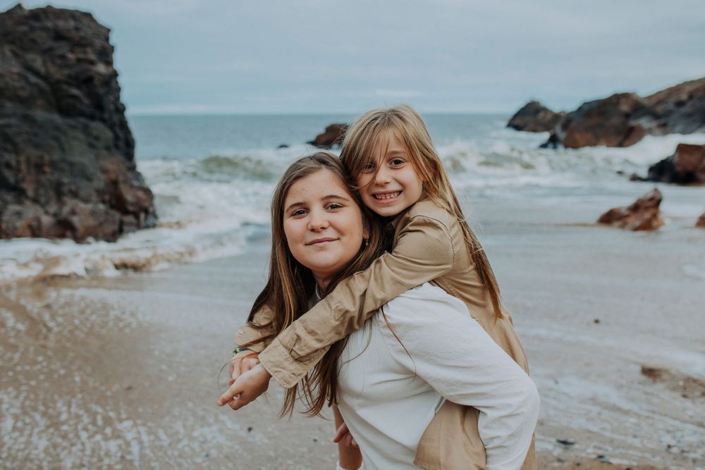 Two friends share a playful piggyback ride on a rocky beach with crashing waves and cloudy skies in the background.