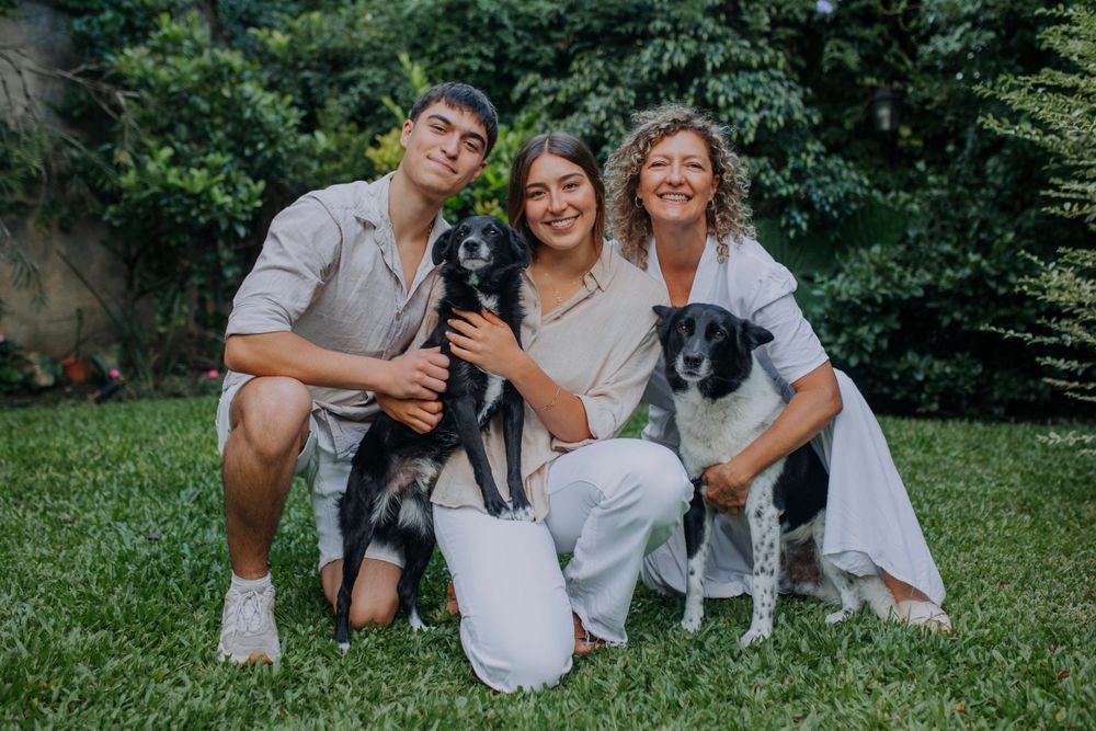 Three people sit on grass with their two black and white dogs in an outdoor garden setting.