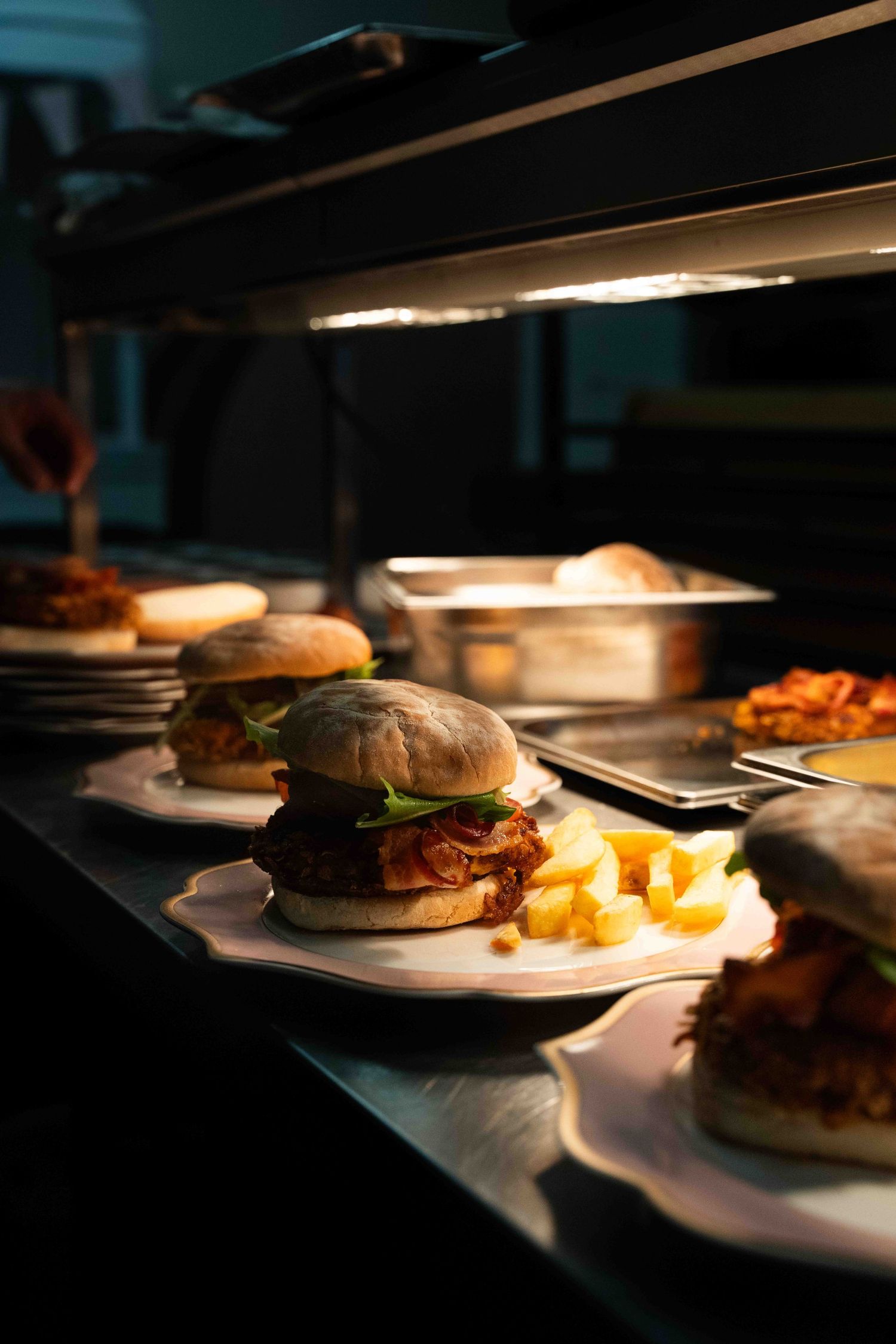 Gourmet burgers with fries on white plates under warm kitchen lighting.