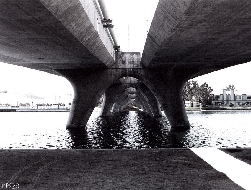 Dramatic perspective underneath a concrete bridge with water reflections in black and white.
