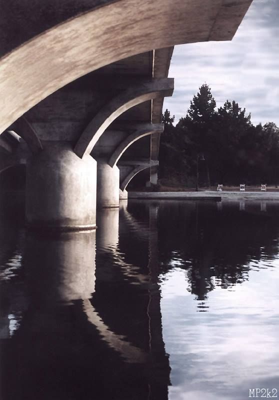 Curved concrete bridge supports casting reflections on still water at sunset.