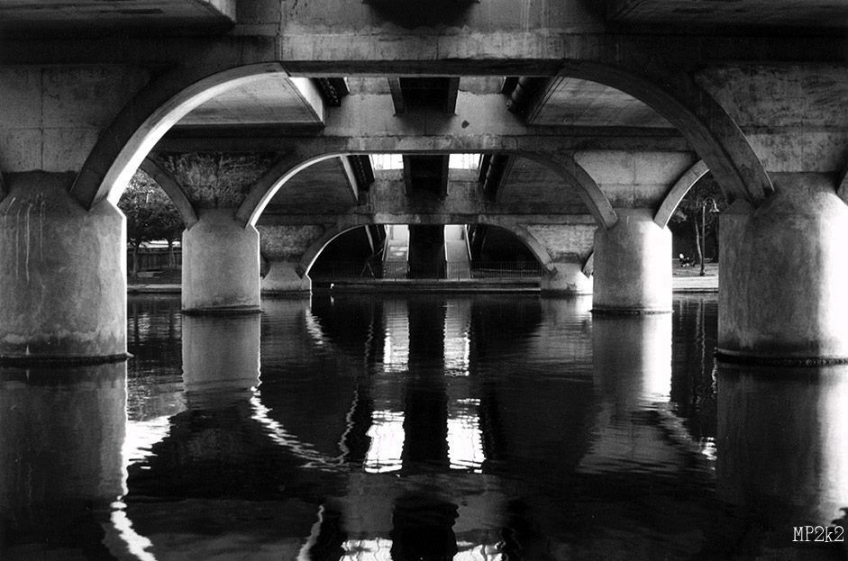 Artistic black and white photo of bridge arches creating symmetrical reflections in water.