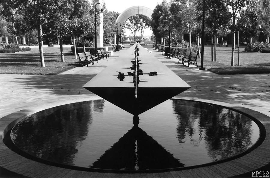 Symmetrical park walkway with reflecting pool and trees leading to curved archway.