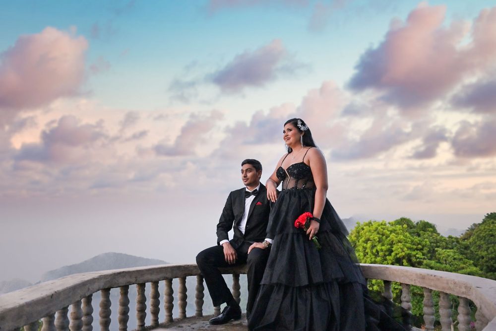 Elegant couple in black formal wear poses on stone balcony at sunset.