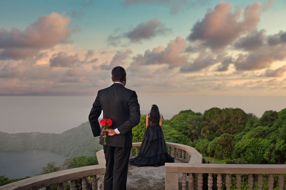 Couple in formal attire stands on hilltop balcony overlooking scenic landscape at sunset.