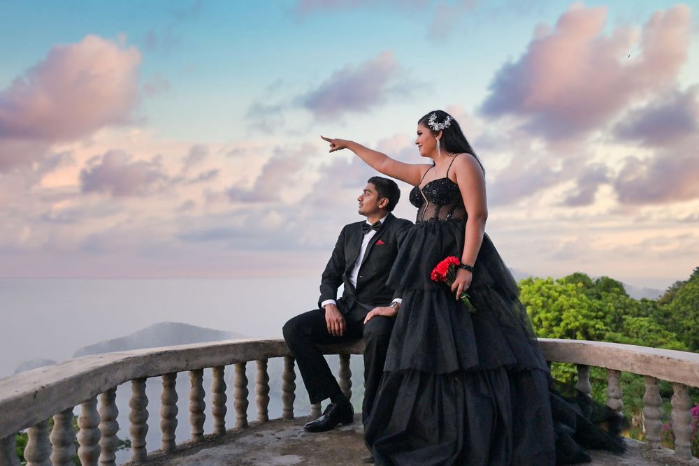 Couple in black formal wear poses dramatically on stone balcony with mountain views.